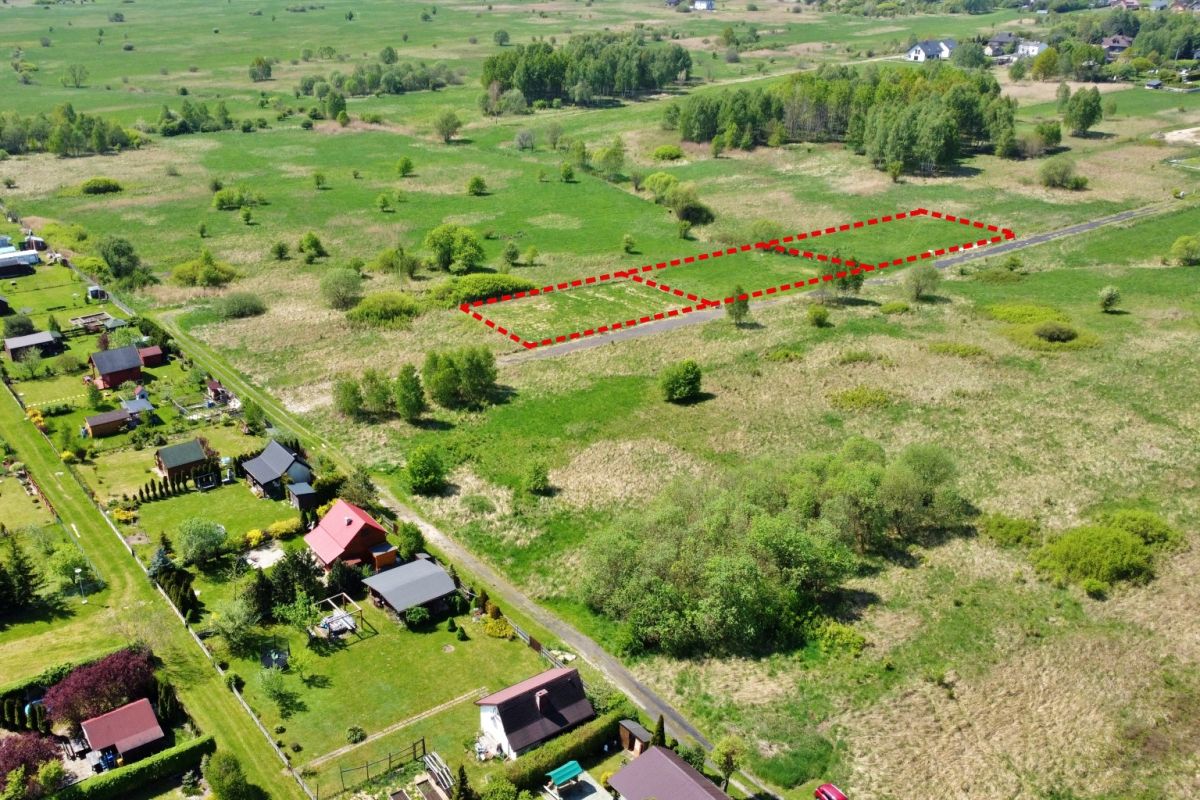 Aerial view of two adjacent vacant lots outlined in red beside a road in a rural area, with green fields and houses nearby.