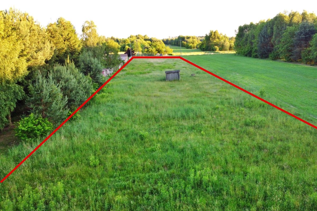 Aerial view of a green field with red boundary lines marking a triangular plot, small shed near center, trees along left and right edges.