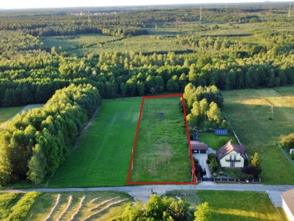 Aerial view of a rectangular grassy plot outlined in red, bordered by trees, with a house and driveway to the right and a road in the foreground.