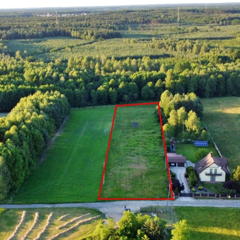 Aerial view of a rectangular grassy plot outlined in red, bordered by trees, with a house and driveway to the right and a road in the foreground.