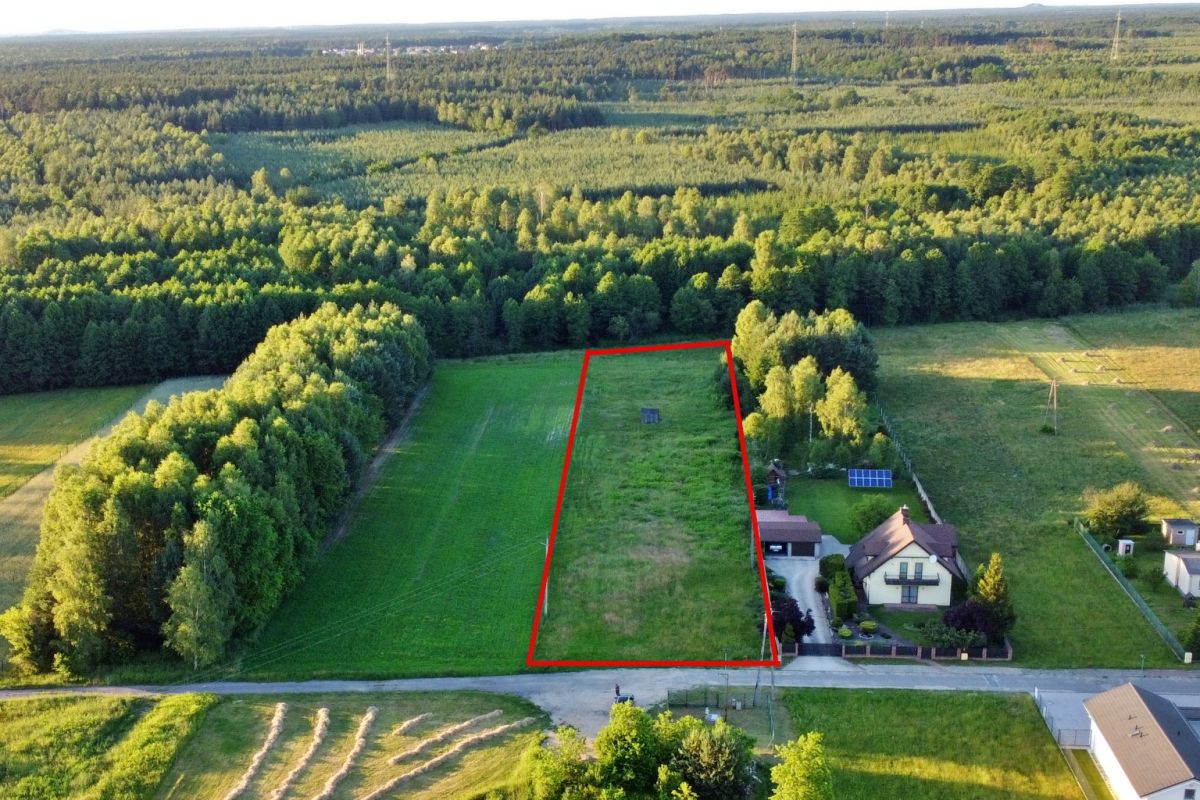 Aerial view of a rectangular grassy plot outlined in red, bordered by trees, with a house and driveway to the right and a road in the foreground.