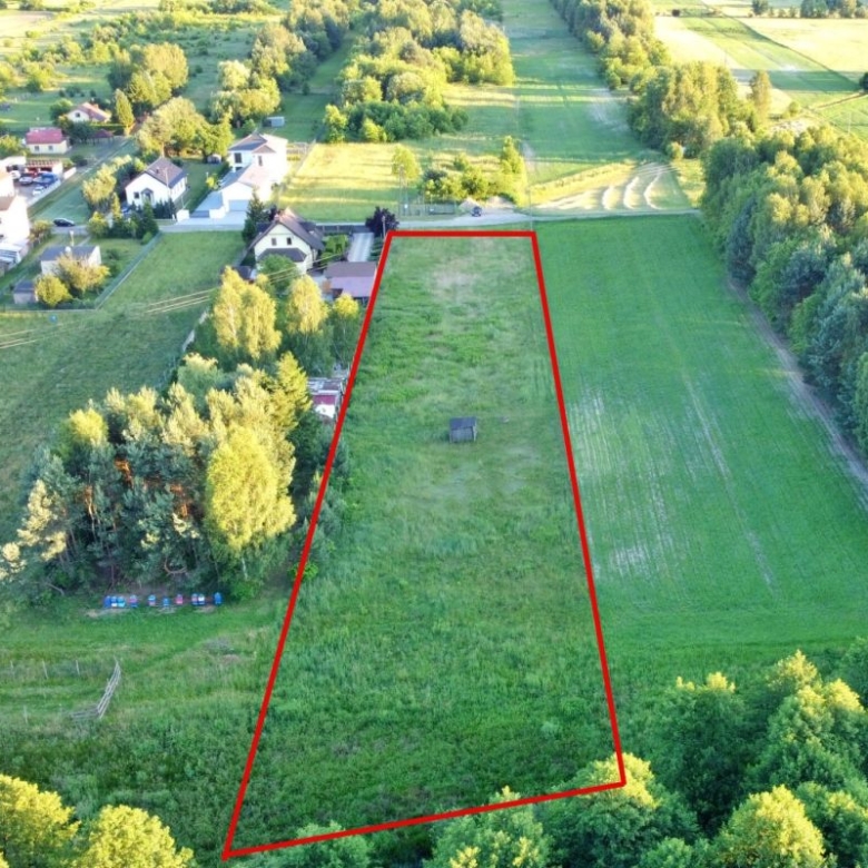 Aerial view of a vacant trapezoid-shaped lot outlined in red, bordered by trees and fields, with a small shed near the center.
