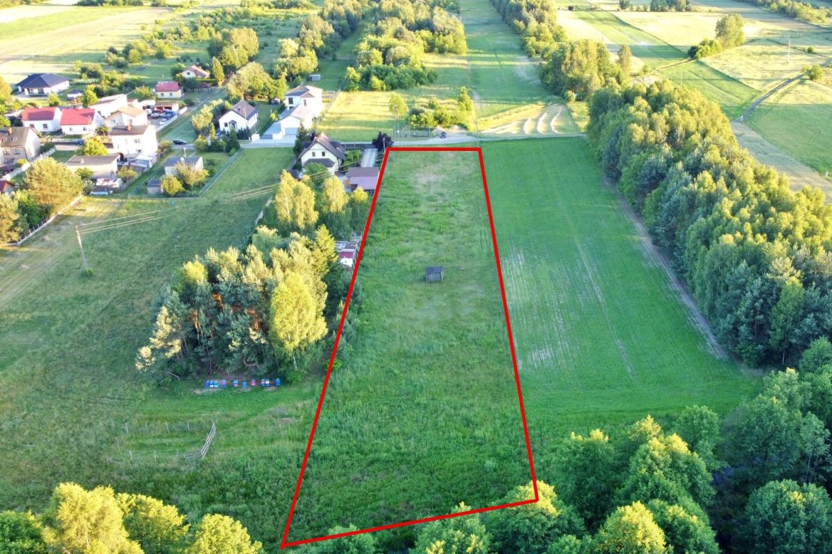 Aerial view of a vacant trapezoid-shaped lot outlined in red, bordered by trees and fields, with a small shed near the center.