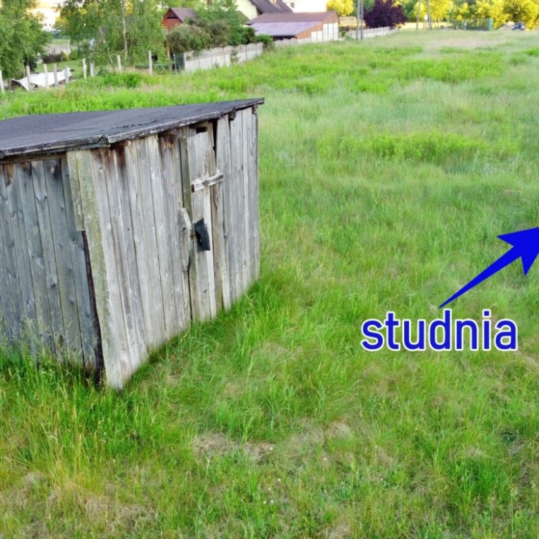 Grassy rural yard with a weathered wooden shed on the left and a round well cap in the foreground on the right.