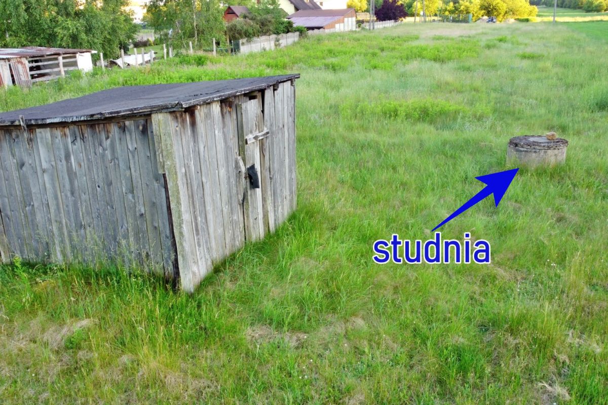 Grassy rural yard with a weathered wooden shed on the left and a round well cap in the foreground on the right.