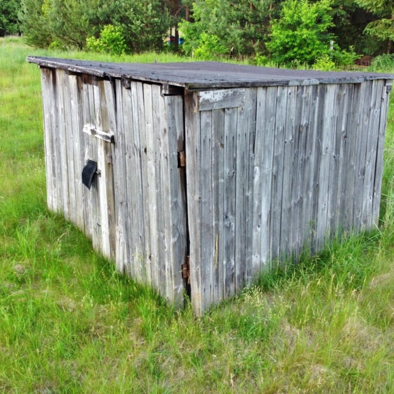 Weathered wooden shed in a grassy field, with a flat, dark roof and a black latch on the door edges nearby trees in the background.