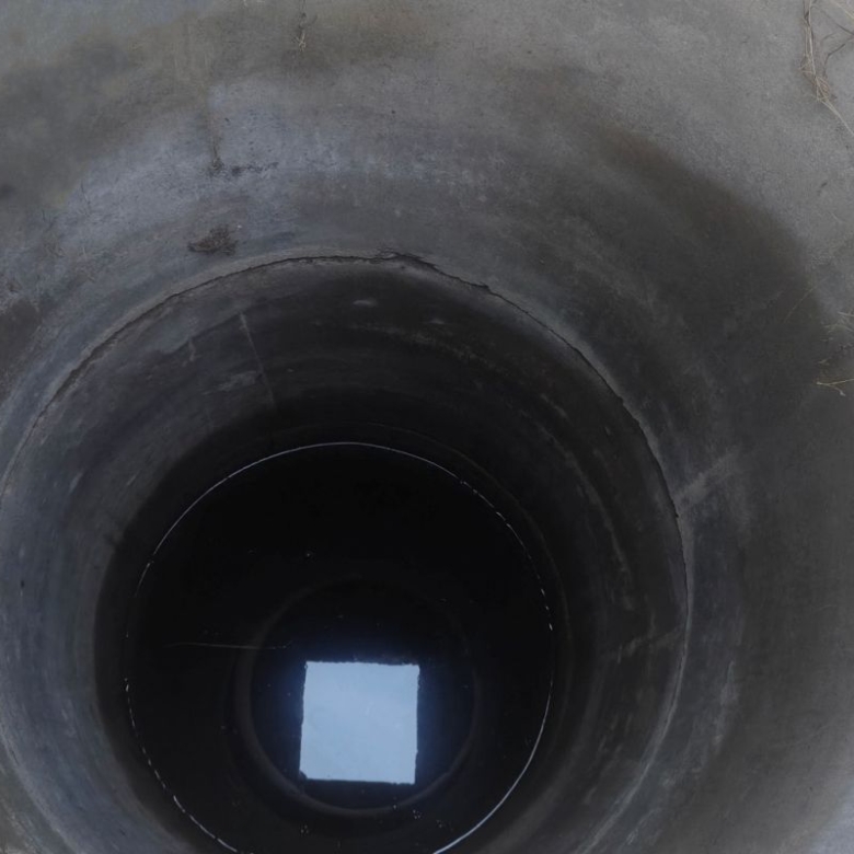 Open vertical concrete well or manhole viewed from above, with dark inner shaft and a bright square of light at the bottom.