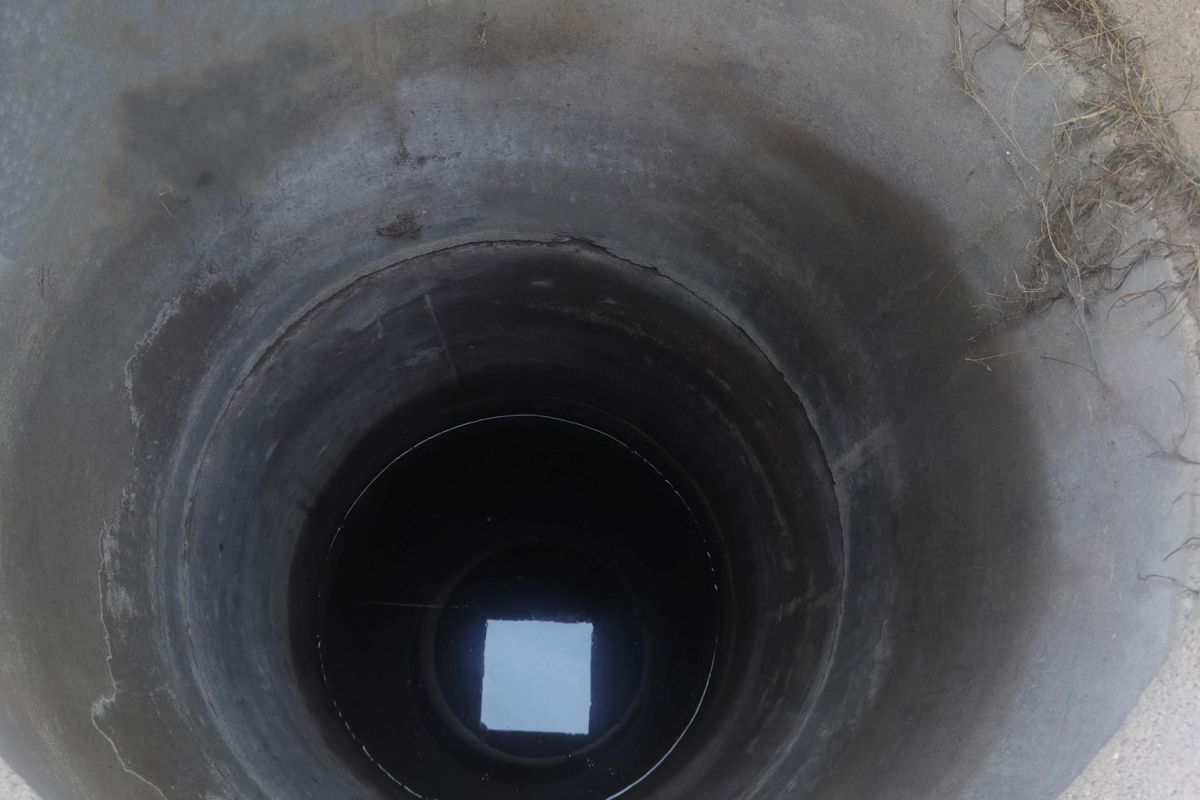 Open vertical concrete well or manhole viewed from above, with dark inner shaft and a bright square of light at the bottom.