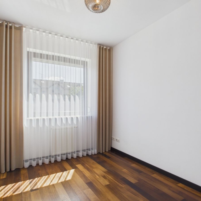 Empty white-walled room with a dark wood floor and a large window dressed in sheer white curtains under beige drapes, sunlight casting stripes on the floor.