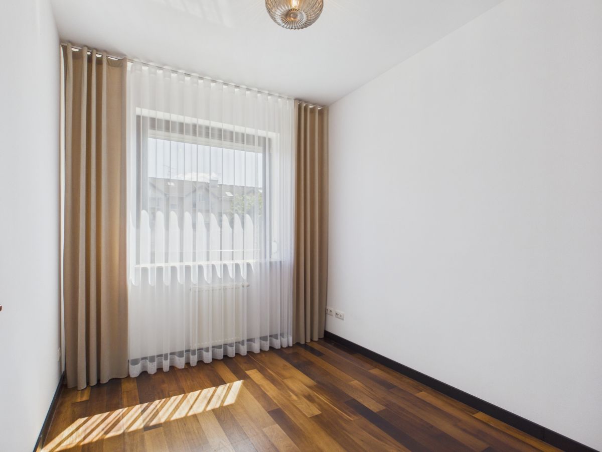 Empty white-walled room with a dark wood floor and a large window dressed in sheer white curtains under beige drapes, sunlight casting stripes on the floor.