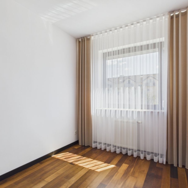 Empty small room with white walls, wooden floor, and a large window with sheer white blinds and beige curtains, sunlight on the floor
