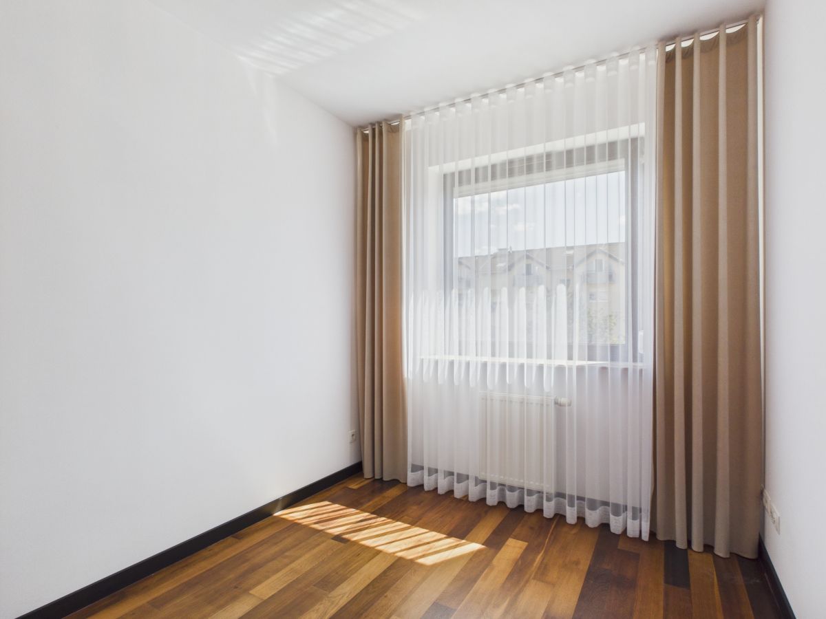 Empty small room with white walls, wooden floor, and a large window with sheer white blinds and beige curtains, sunlight on the floor