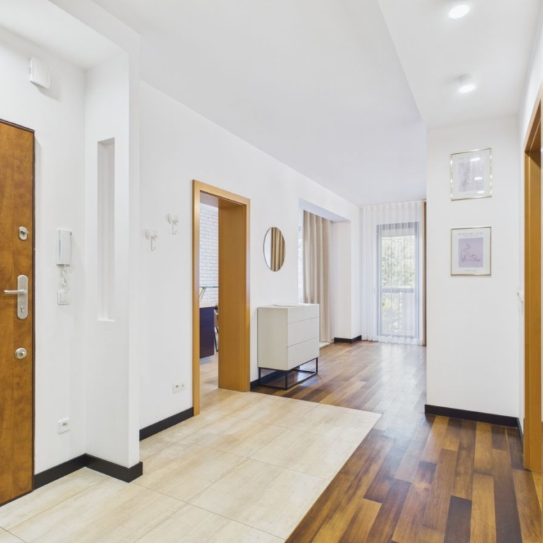 Bright hallway in a contemporary home with a wooden front door on the left, white walls, and a doorway opening to a living area; mix of tile and wood flooring. In the middle a white dresser and a round mirror on the wall.