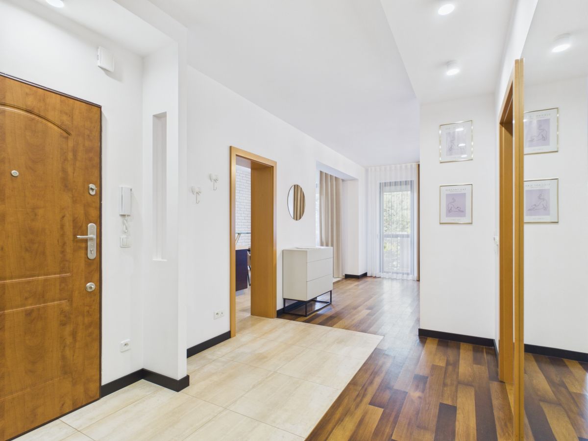 Bright hallway in a contemporary home with a wooden front door on the left, white walls, and a doorway opening to a living area; mix of tile and wood flooring. In the middle a white dresser and a round mirror on the wall.