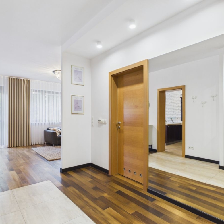 Bright, modern foyer with dark wood flooring, a large wall mirror, and a wooden door; living area with sheer curtains and a sofa visible in the background.