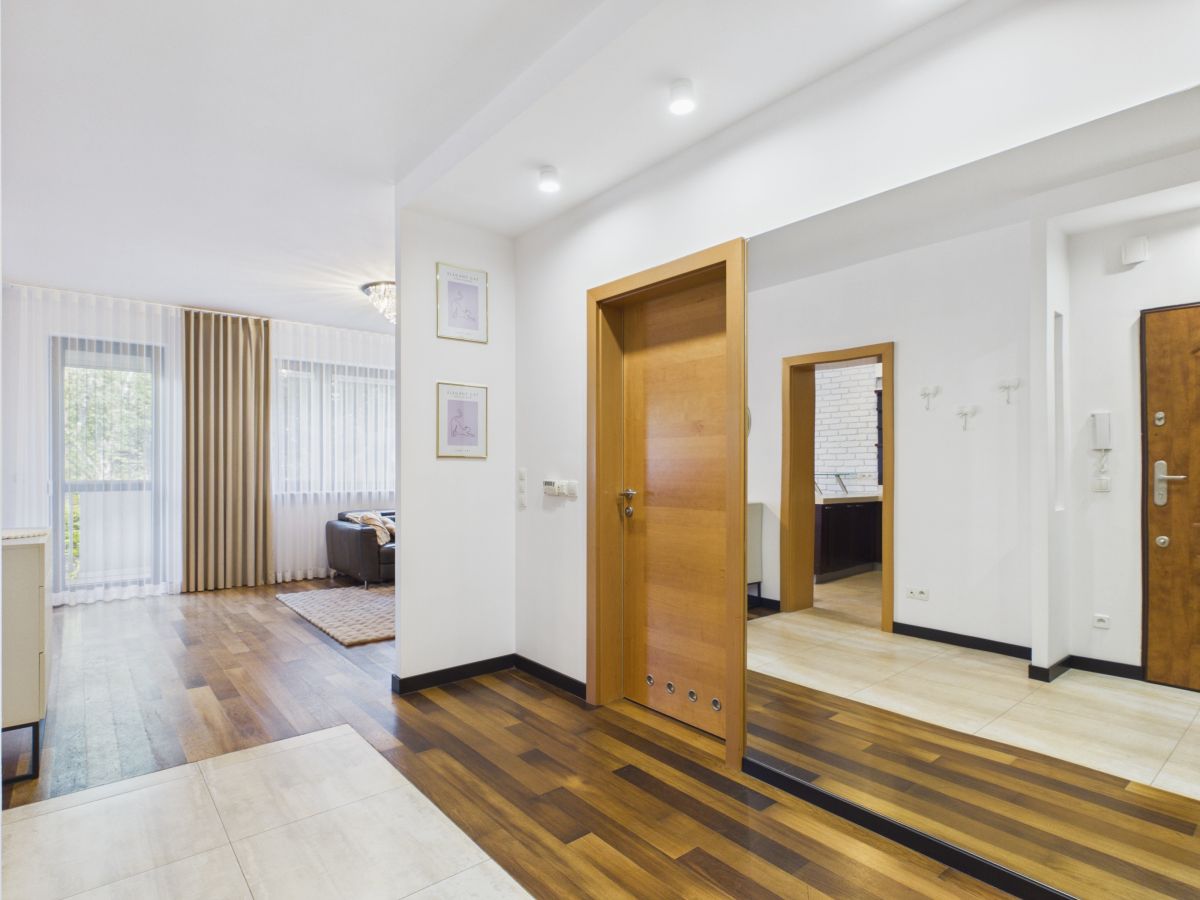 Bright, modern foyer with dark wood flooring, a large wall mirror, and a wooden door; living area with sheer curtains and a sofa visible in the background.
