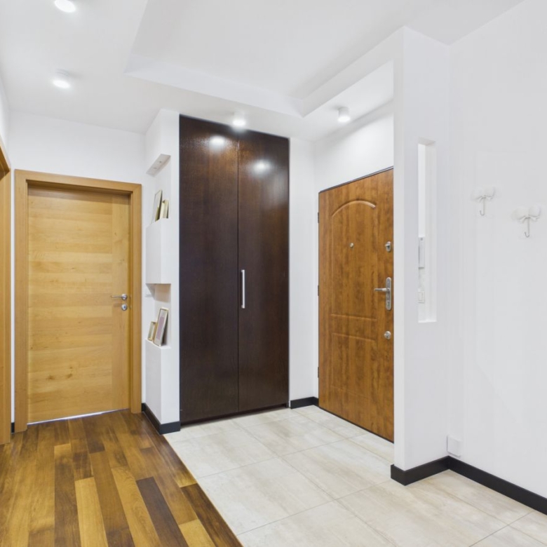 Modern hallway interior with three wooden doors (two lighter, one dark) and a tall dark wardrobe; light tiled floor and white walls.