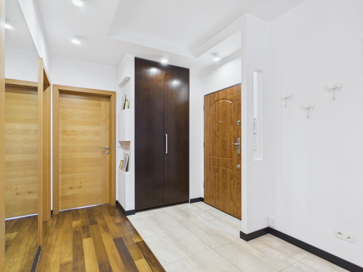 Modern hallway interior with three wooden doors (two lighter, one dark) and a tall dark wardrobe; light tiled floor and white walls.
