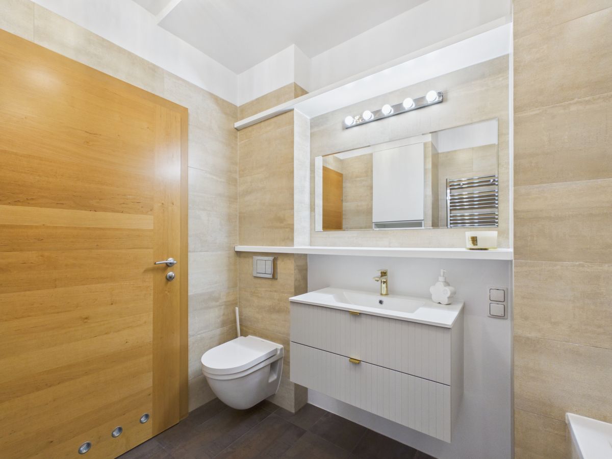 Modern bathroom with wall-mounted toilet, white vanity, large mirror, and beige tile walls in a bright space.