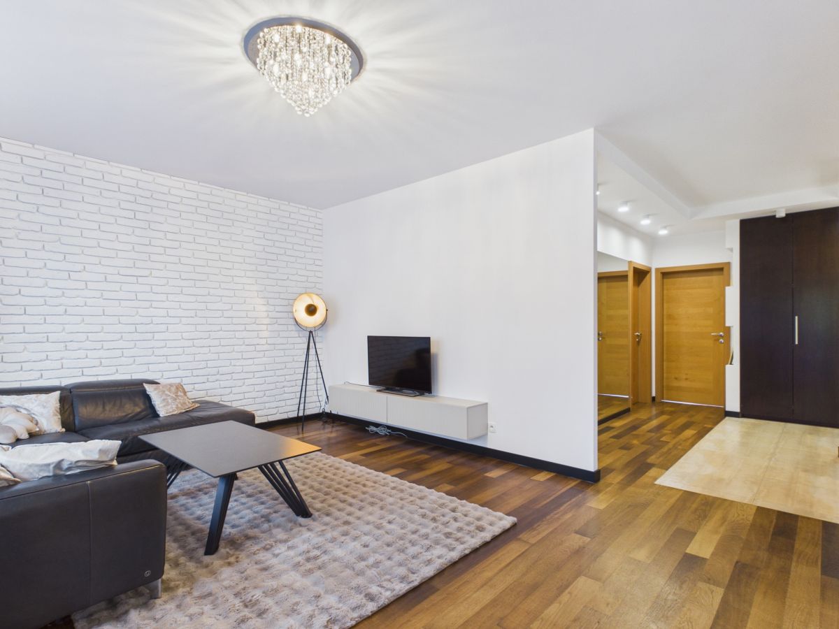 Modern living room with white brick accent wall, chandelier, dark leather sofa, and a rectangular coffee table on a light rug.