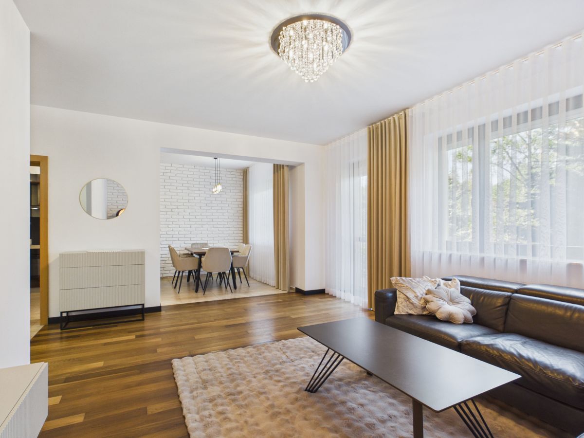Bright, modern living room with a dark leather sofa, rectangular coffee table, and a cream rug by large windows with sheer curtains.