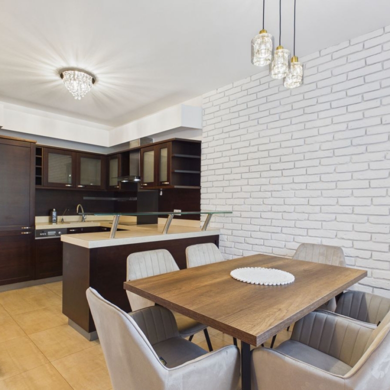 Open-concept kitchen and dining area with white brick wall, dark wood cabinets, and a wooden dining table with six upholstered chairs.