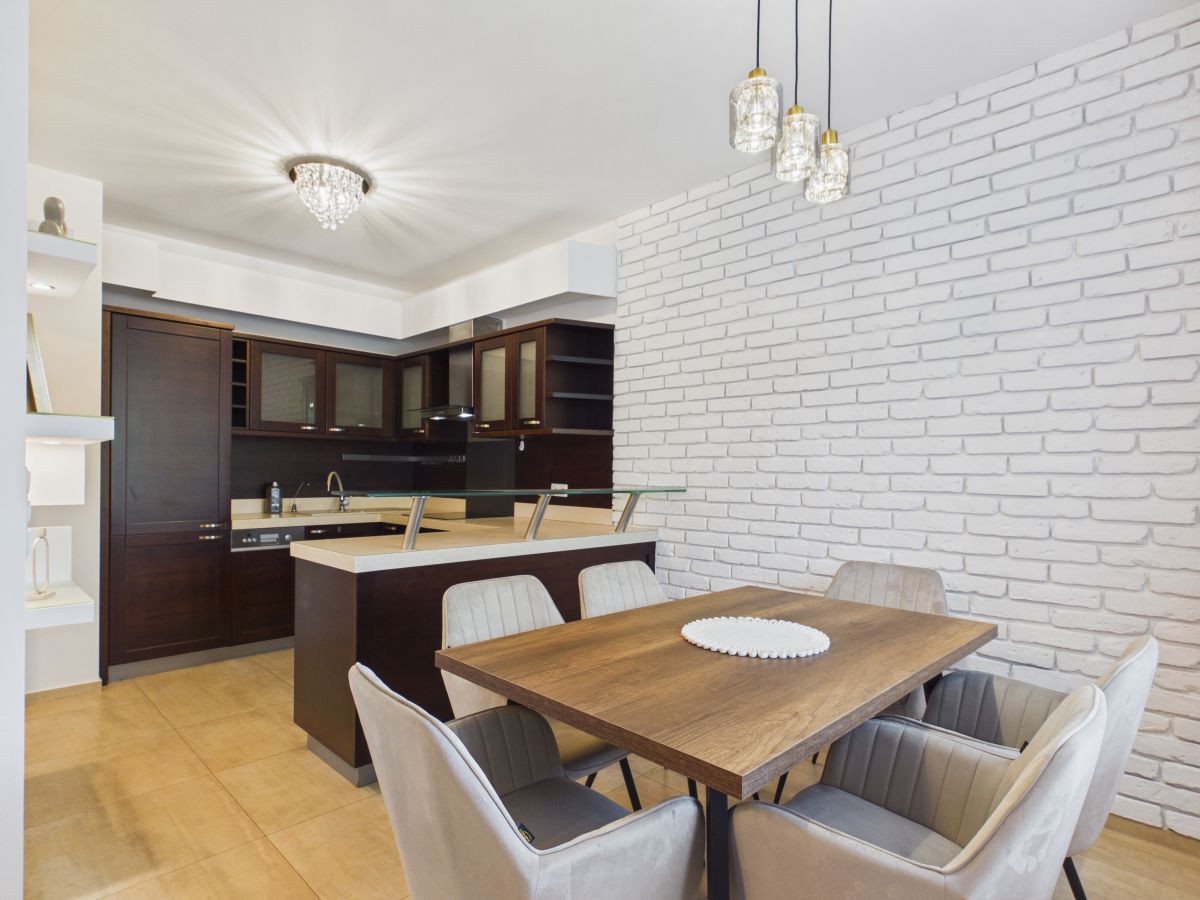 Open-concept kitchen and dining area with white brick wall, dark wood cabinets, and a wooden dining table with six upholstered chairs.