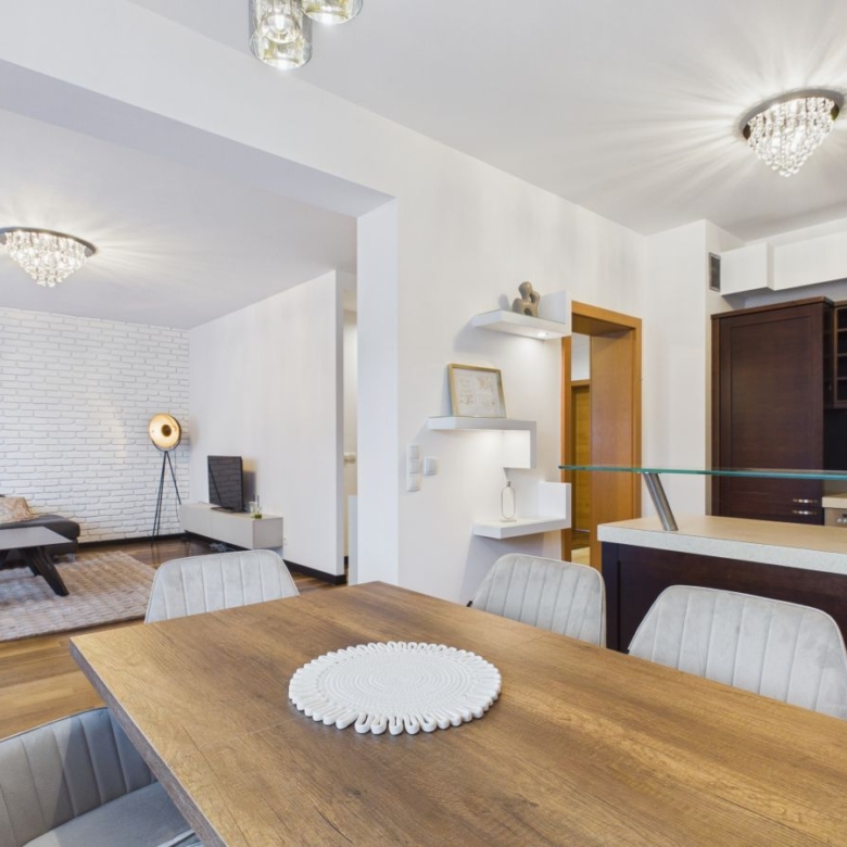 Open-concept living and dining area with a wooden table, gray upholstered chairs, white walls, and a brick accent wall in the living room.