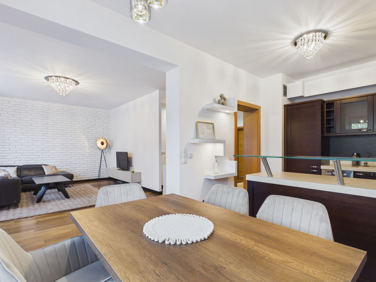 Open-concept living and dining area with a wooden table, gray upholstered chairs, white walls, and a brick accent wall in the living room.