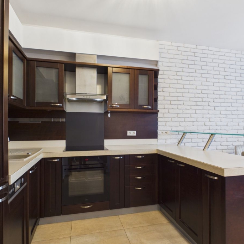 Modern L-shaped kitchen with dark wood cabinets, beige countertops, stainless range hood, and glass-railed island in a bright space.