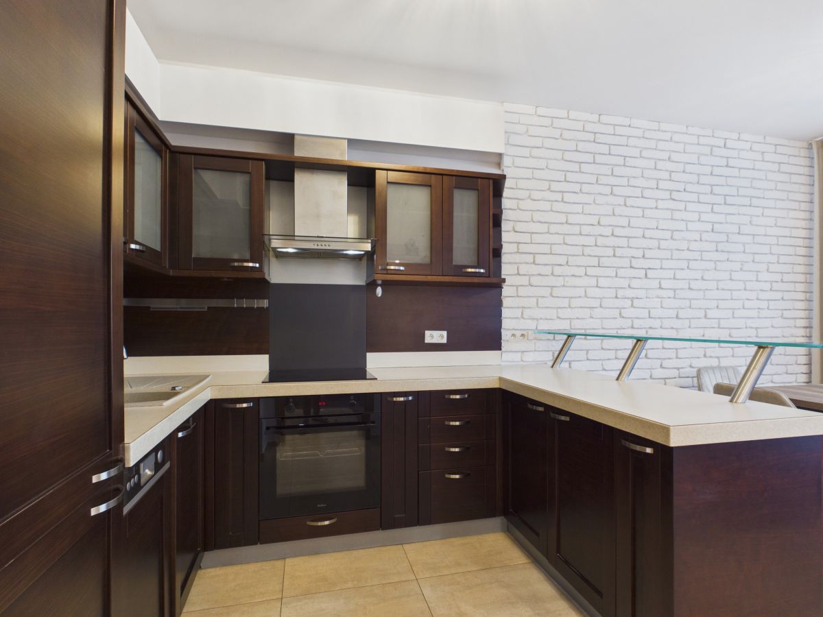 Modern L-shaped kitchen with dark wood cabinets, beige countertops, stainless range hood, and glass-railed island in a bright space.