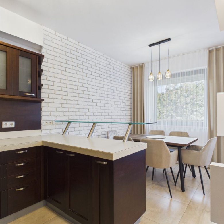 Modern open-plan kitchen and dining area with dark wood cabinets, white brick accent wall, and a glass-top island beside a dining table with beige chairs.