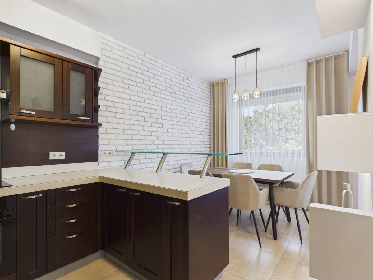 Modern open-plan kitchen and dining area with dark wood cabinets, white brick accent wall, and a glass-top island beside a dining table with beige chairs.