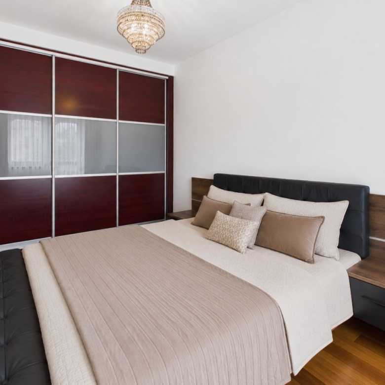 Bedroom with a queen bed, beige bedding and multiple throw pillows, dark wood headboard and matching side tables, and a large sliding wardrobe with frosted panels.