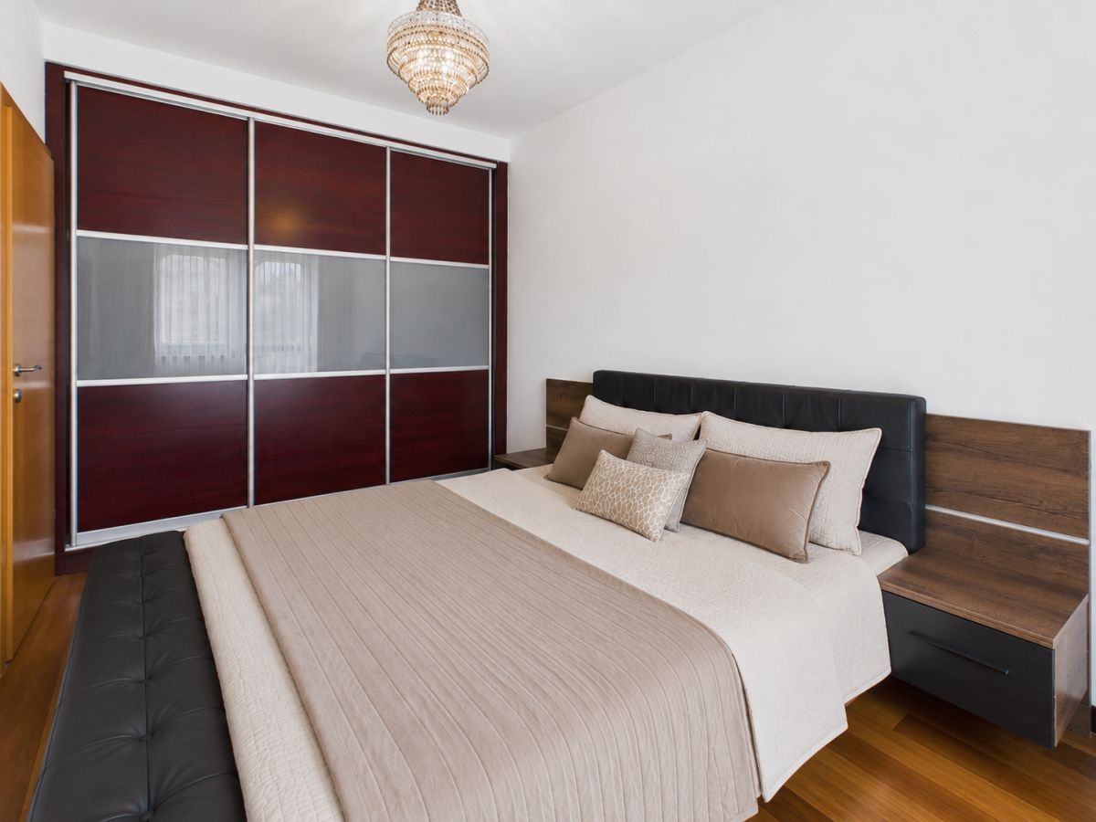 Bedroom with a queen bed, beige bedding and multiple throw pillows, dark wood headboard and matching side tables, and a large sliding wardrobe with frosted panels.