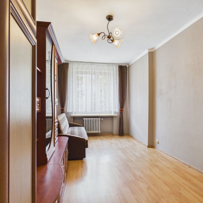 Narrow living room with light hardwood floor, a brown sofa and tall wooden cabinet along the left wall, and a large window with sheer white curtains and brown drapes behind.