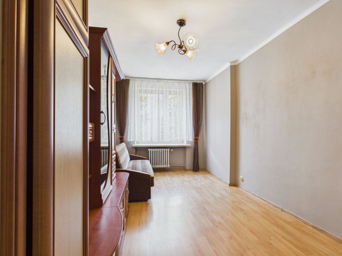 Narrow living room with light hardwood floor, a brown sofa and tall wooden cabinet along the left wall, and a large window with sheer white curtains and brown drapes behind.