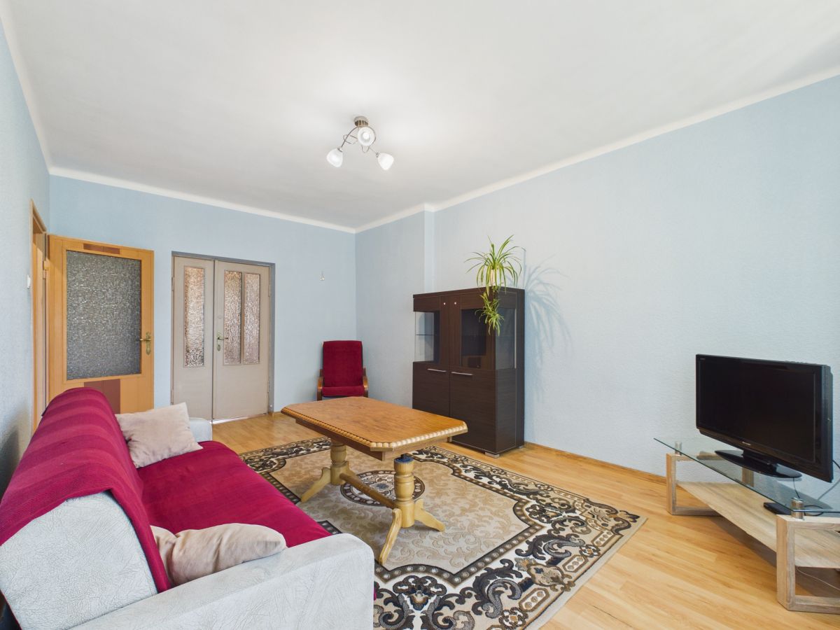 Living room with light blue walls, red sofa, wooden coffee table, patterned area rug, and TV on a glass stand opposite the sofa.