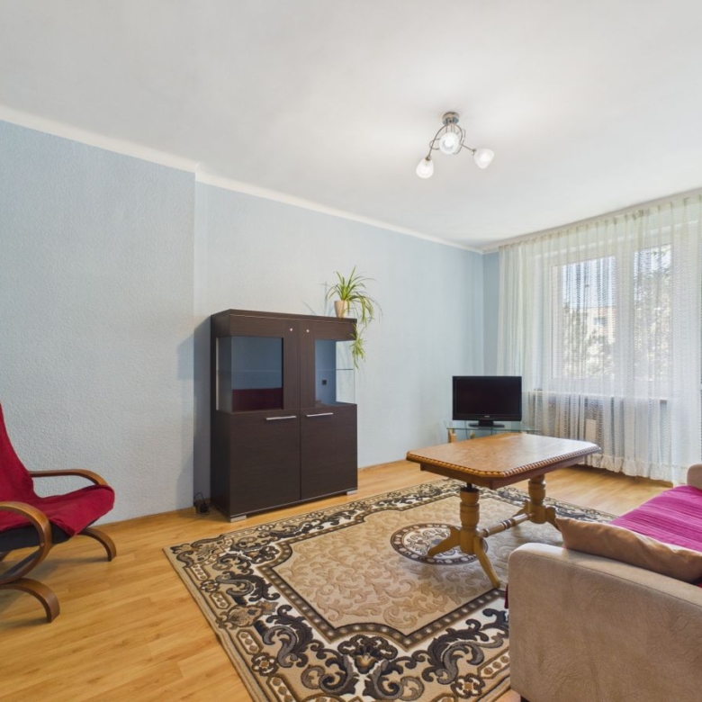 Living room with light blue walls, beige sofa with red throw, wooden coffee table, and a dark cabinet near a large window with sheer curtains.