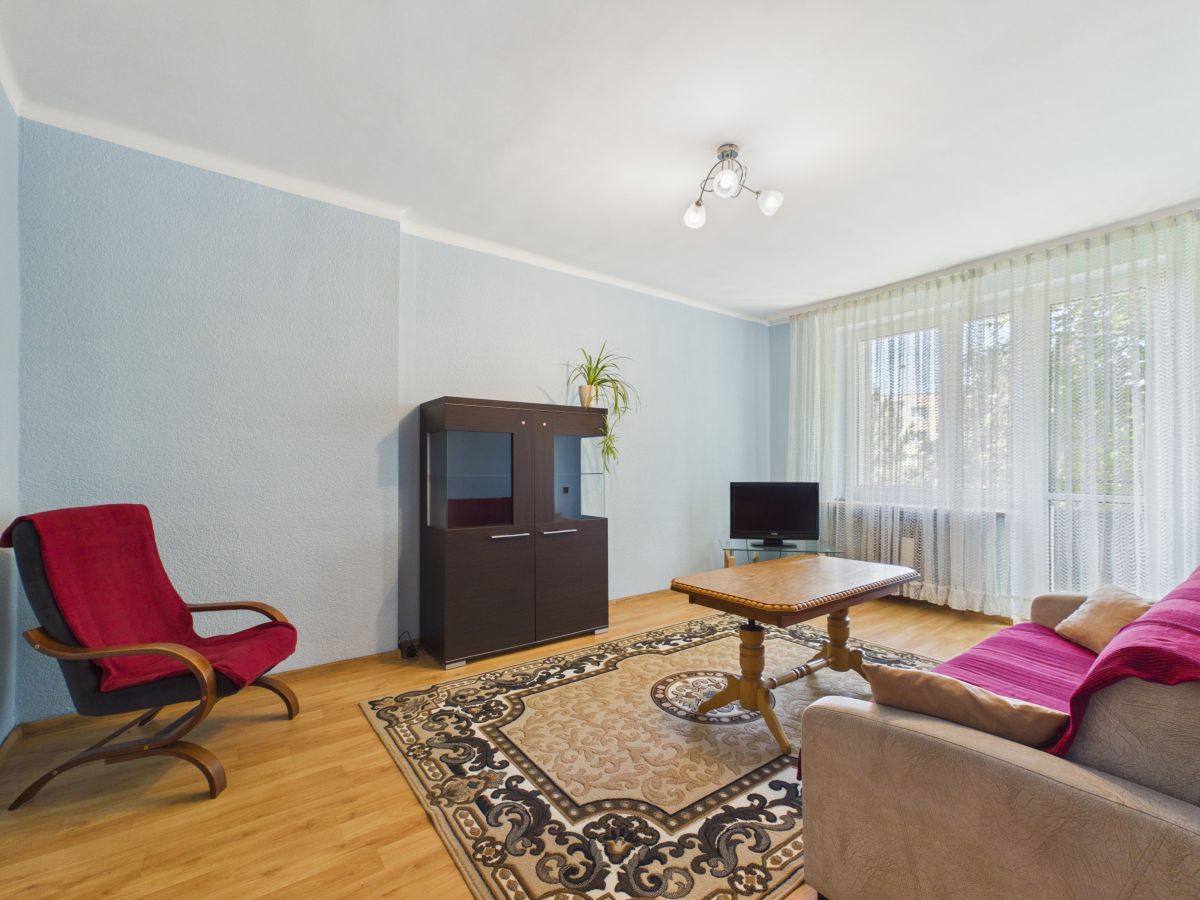Living room with light blue walls, beige sofa with red throw, wooden coffee table, and a dark cabinet near a large window with sheer curtains.