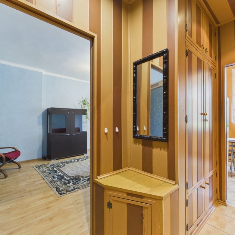 Wood-paneled hallway with built-in cupboards and a black-framed wall mirror. Left opens into a blue-walled living area with a red chair and cabinet, rug, and wood floor. Right leads to a tiled patio/kitchen area with a table and chairs.
