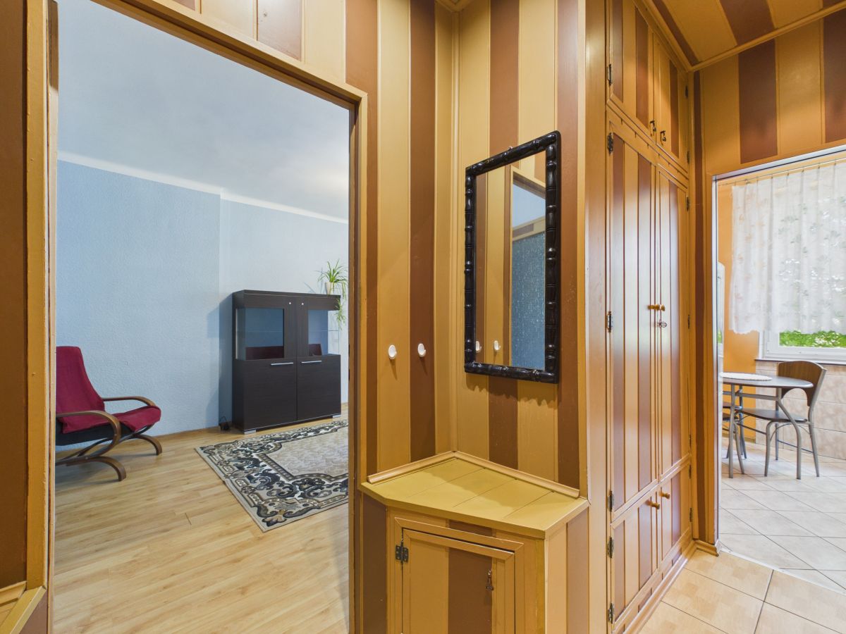 Wood-paneled hallway with built-in cupboards and a black-framed wall mirror. Left opens into a blue-walled living area with a red chair and cabinet, rug, and wood floor. Right leads to a tiled patio/kitchen area with a table and chairs.