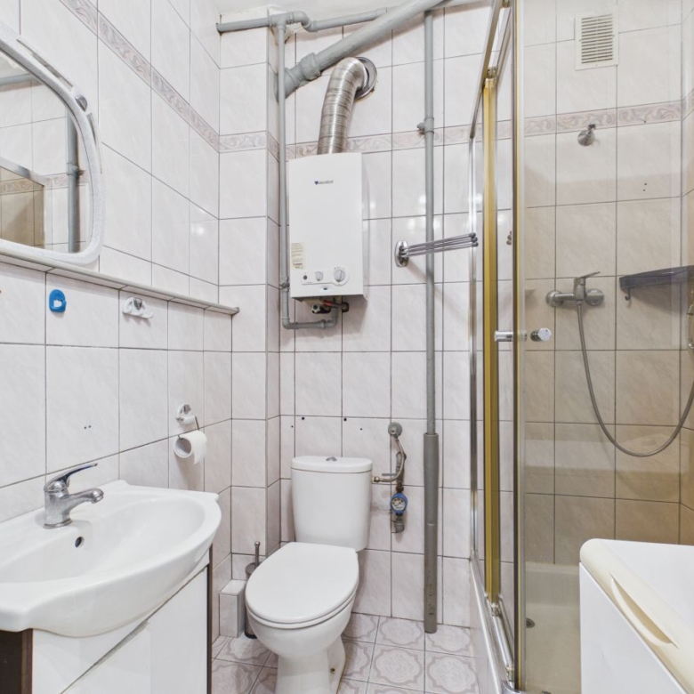 Small tiled bathroom with a sink, toilet, and a glass-enclosed shower; a boiler and exposed pipes run along the walls above the toilet.