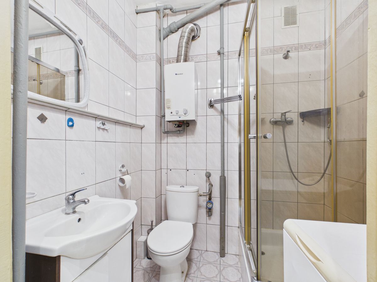 Small tiled bathroom with a sink, toilet, and a glass-enclosed shower; a boiler and exposed pipes run along the walls above the toilet.