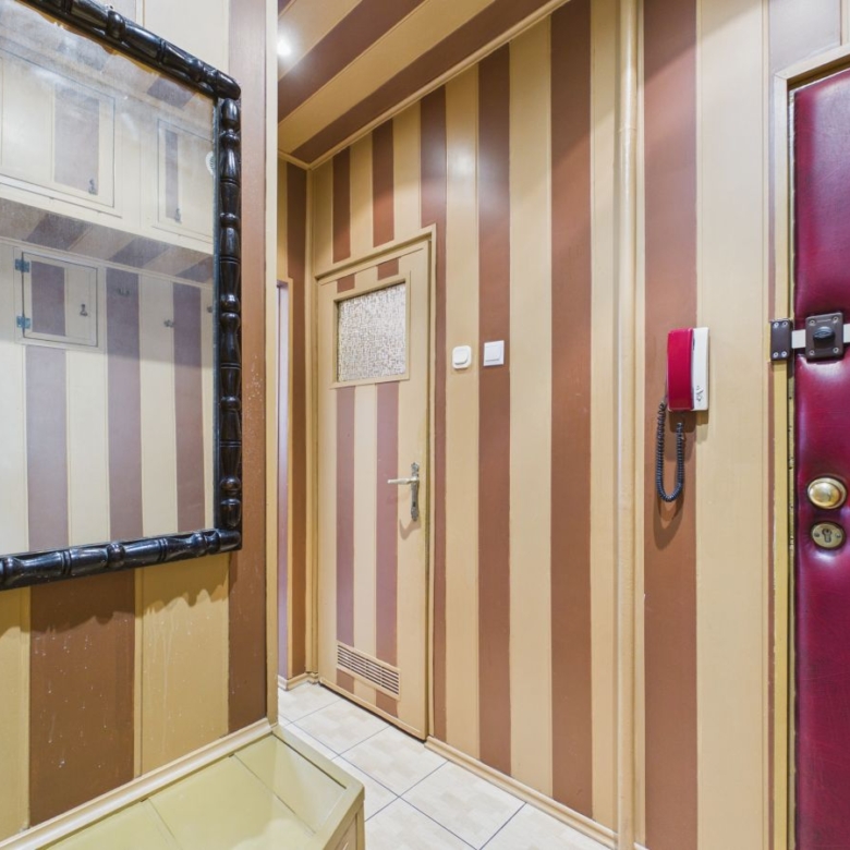 Narrow striped hallway with brown-beige walls, a door with a small window, a red wall intercom, and a padded burgundy door to the right, plus a large black-framed mirror on the left.