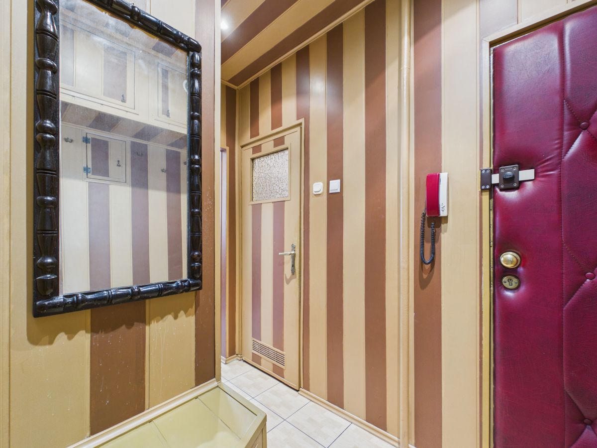 Narrow striped hallway with brown-beige walls, a door with a small window, a red wall intercom, and a padded burgundy door to the right, plus a large black-framed mirror on the left.