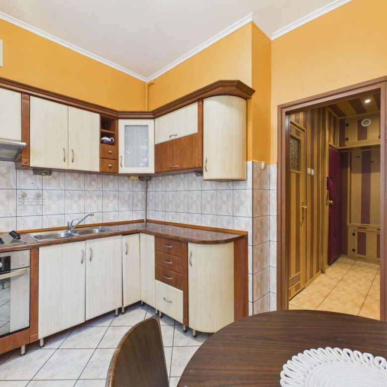 Orange-walled kitchen with light wood cabinets, tiled backsplash, gas stove and oven, stainless sink, and a round dining table in the foreground.