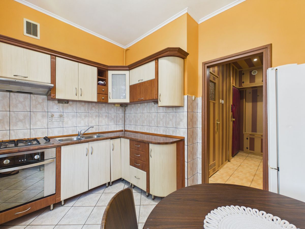 Orange-walled kitchen with light wood cabinets, tiled backsplash, gas stove and oven, stainless sink, and a round dining table in the foreground.