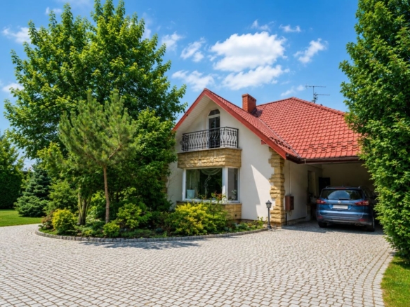 Two-story house with red tiled roof, stone accents, and a driveway bordered by lush green trees under a blue sky.