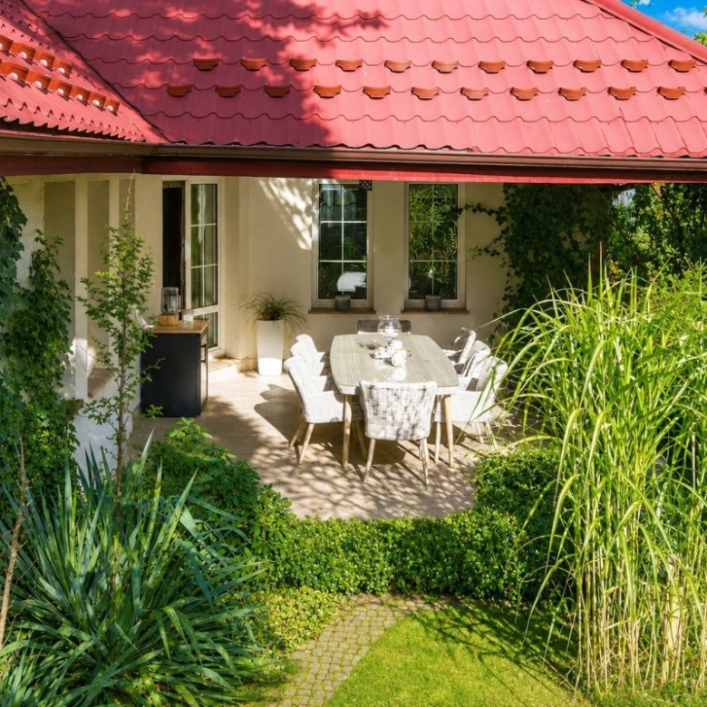 Sunlit patio dining area under a red-tiled roof, with a long table and grey wicker chairs surrounded by lush green plants.